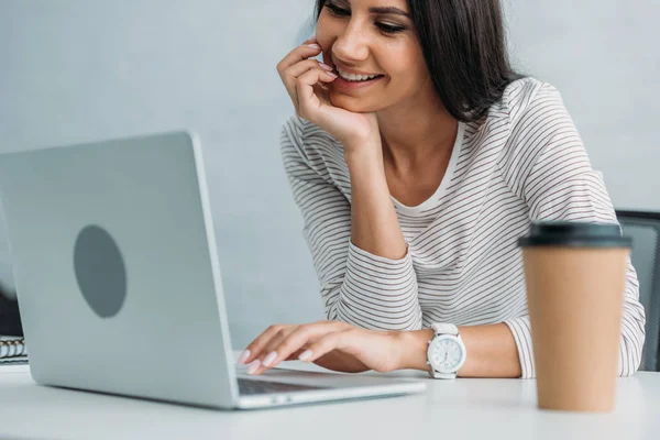 Atractiva y morena mujer sonriendo y utilizando el ordenador portátil en el apartamento - foto de stock