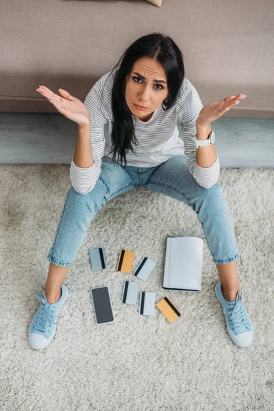 Vista de ángulo alto de la mujer hermosa y triste sentado en el suelo con tarjetas de crédito, portátil y teléfono inteligente - foto de stock