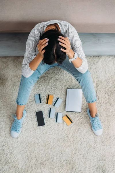 Hochwinkelaufnahme einer aufgebrachten Frau, die mit Kreditkarten, Notebook und Smartphone auf dem Boden sitzt — Stockfoto