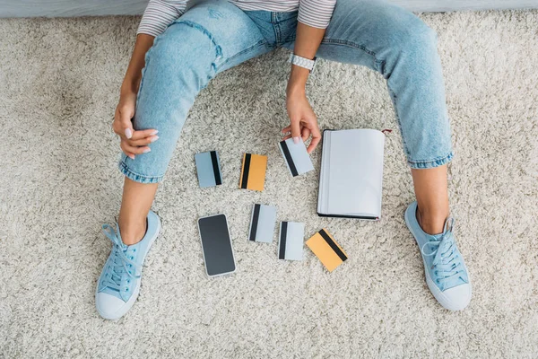 Partial view of  woman sitting on floor with notebook, smartphone and holding credit card — Stock Photo