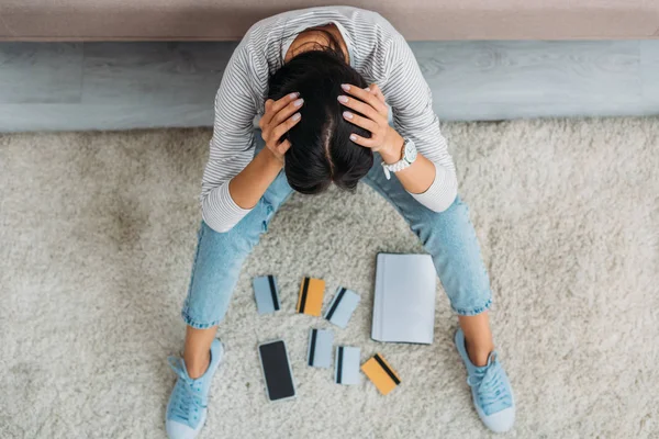 Vista superior de la triste mujer sentada en el suelo con portátil, teléfono inteligente y tarjetas de crédito - foto de stock