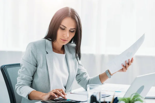 Mulher atraente e morena em desgaste formal segurando documento e usando calculadora — Fotografia de Stock