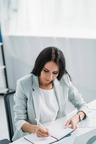 Attraktive und brünette Frau in formeller Kleidung, die in Notizbuch schreibt — Stockfoto