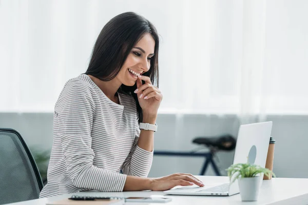 Schöne und brünette Frau, die lächelt und auf den Bildschirm ihres Laptops blickt — Stockfoto