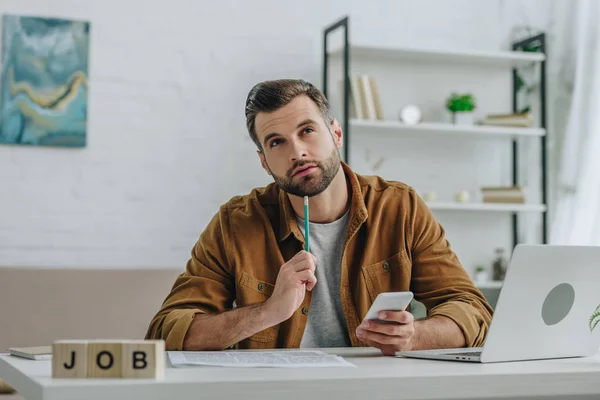 Hombre de ensueño sosteniendo teléfono inteligente, lápiz y mirando hacia arriba entre cubos con trabajo de letras - foto de stock