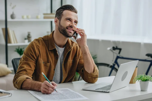 Schöner und lächelnder Mann, der mit Smartphone spricht und Bleistift in der Hand hält — Stockfoto