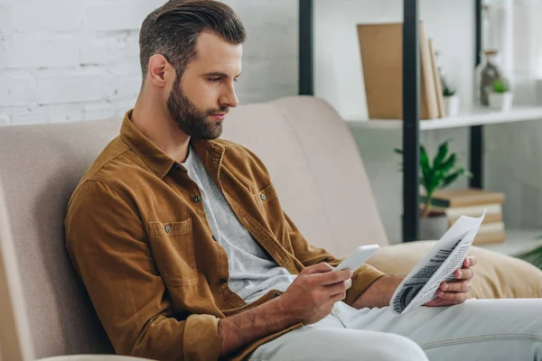 Homem bonito sentado no sofá, usando smartphone e segurando jornal — Fotografia de Stock