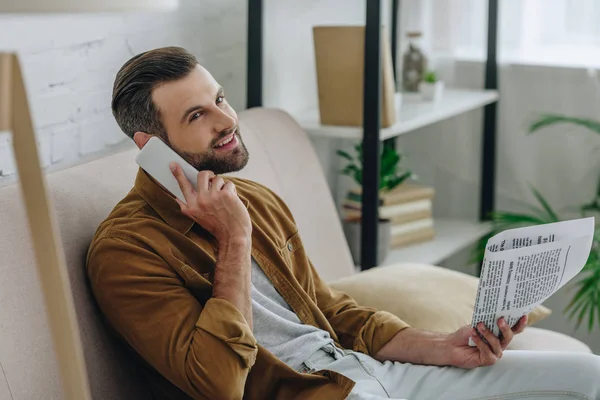 Homem bonito falando no smartphone e segurando jornal no apartamento — Fotografia de Stock