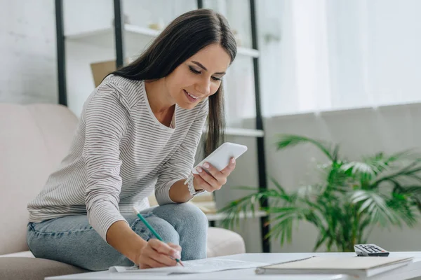 Femme heureuse et attrayante souriant, en utilisant smartphone et crayon tenant — Photo de stock