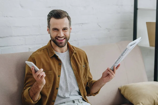 Hombre guapo sosteniendo teléfono inteligente y periódico, sonriendo y mirando a la cámara - foto de stock