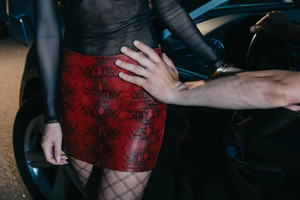 Cropped view of man touching seductive prostitute in red skirt standing near car — Stock Photo