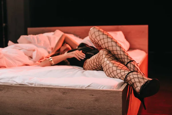 Selective focus of sexy woman in mesh stockings and heels lying on bed on black — Stock Photo