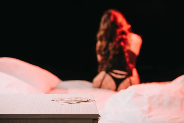 Селективный фокус долларовых банкнот на прикроватной тумбочке рядом с сексуальной женщиной, сидящей на кровати изолированной на черном — стоковое фото