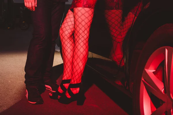Vista ritagliata della donna in gonna rossa e calze a rete in piedi con l'uomo vicino auto — Foto stock