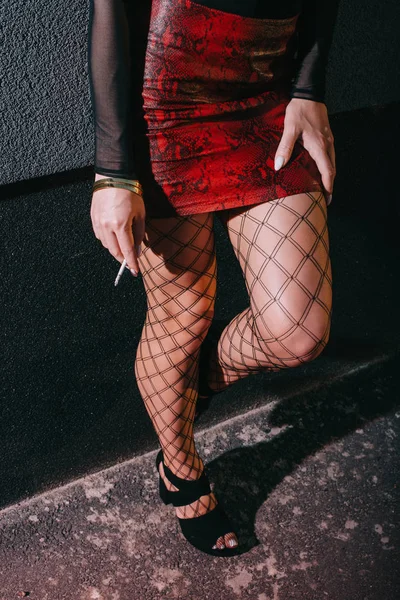 Recortado vista de sexy prostituta en rojo falda de pie cerca de la pared con cigarrillo - foto de stock