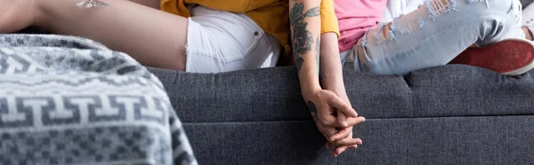 Панорамный снимок двух лесбиянок, держащихся за руки, сидящих на диване в гостиной — стоковое фото
