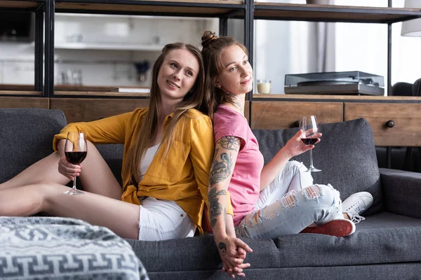 Две улыбающиеся лесбиянки держатся за бокалы и держатся за руки, сидя на диване в гостиной — стоковое фото