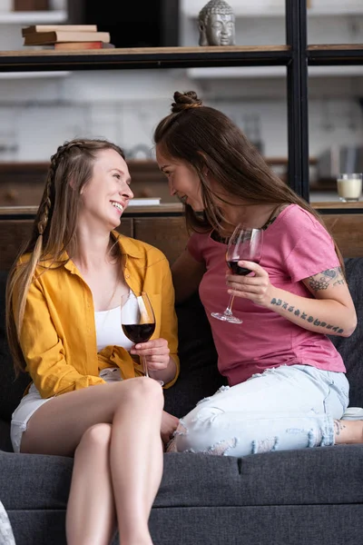 Deux lesbiennes souriantes tenant des verres à vin et se regardant assis sur le canapé dans le salon — Photo de stock