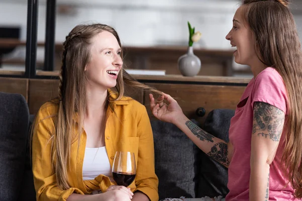 Sorrindo lésbica segurando copo de vinho e olhando para a namorada enquanto sentado no sofá na sala de estar — Fotografia de Stock