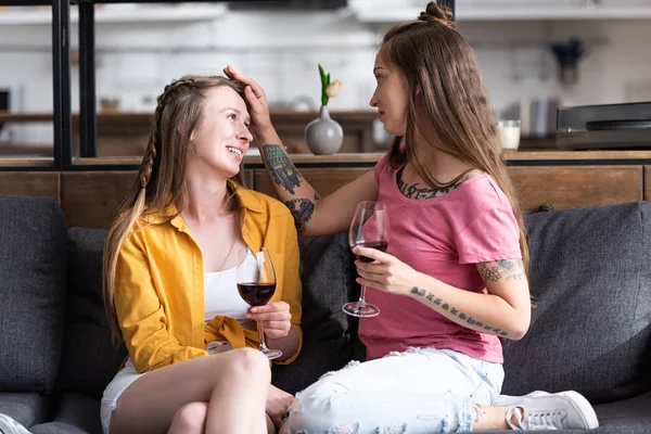 Duas lésbicas segurando copos de vinho e olhando um para o outro enquanto sentado no sofá na sala de estar — Fotografia de Stock