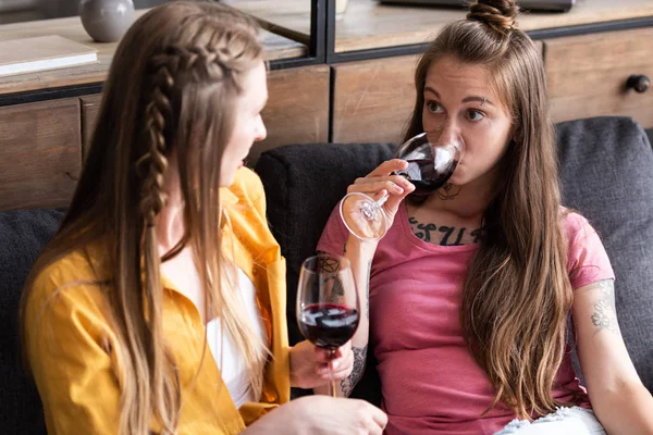 Duas lésbicas segurando copos de vinho e olhando um para o outro enquanto sentado no sofá na sala de estar — Fotografia de Stock