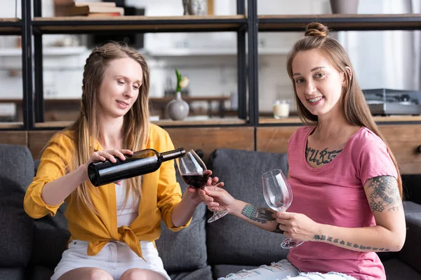 Two lesbians holding wine glasses and pouring wine while sitting on sofa in living room — Stock Photo
