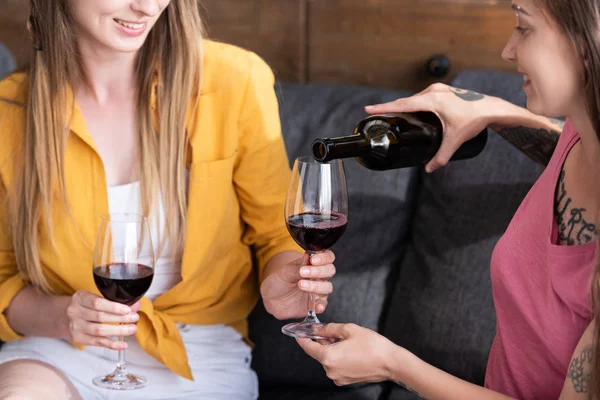 Vista parcial de dos lesbianas sosteniendo copas de vino y vertiendo vino mientras están sentadas en el sofá en la sala de estar - foto de stock