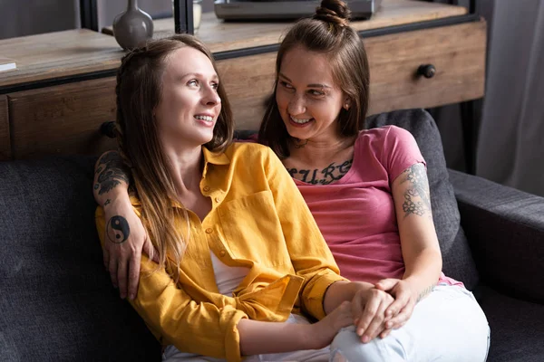 Zwei hübsche Lesben umarmen sich, während sie auf dem Sofa im Wohnzimmer sitzen — Stockfoto