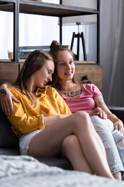 Dos hermosas lesbianas abrazando mientras se sienta en el sofá en la sala de estar - foto de stock