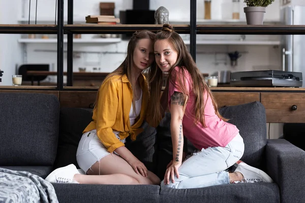 Duas lésbicas sorridentes sentadas no sofá e olhando para a câmera na sala de estar — Fotografia de Stock