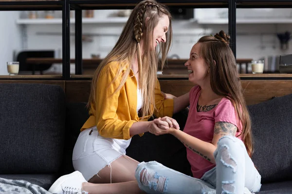 Dos lesbianas sonrientes cogidas de la mano mientras se sientan en el sofá en la sala de estar - foto de stock