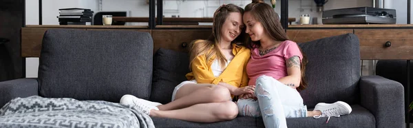 Панорамний знімок двох усміхнених лесбійок, що тримаються за руки, сидячи на дивані у вітальні — стокове фото