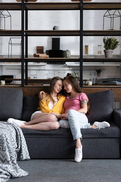 Dos hermosas lesbianas abrazando mientras se sienta en el sofá en la sala de estar - foto de stock