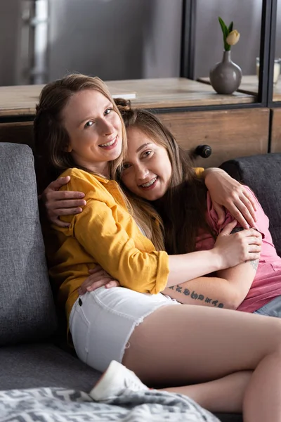 Две симпатичные лесбиянки обнимаются, сидя на диване в гостиной — стоковое фото