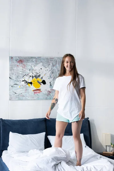 Ganzkörperansicht des glücklichen tätowierten Mädchens, das auf dem Bett im Schlafzimmer steht — Stockfoto