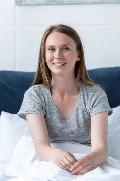 Jolie femme souriante assise sous la couverture sur le lit et regardant la caméra — Photo de stock