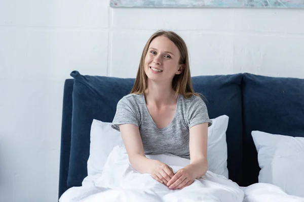 Attraktive lächelnde Frau sitzt unter Decke auf dem Bett und blickt in die Kamera — Stockfoto