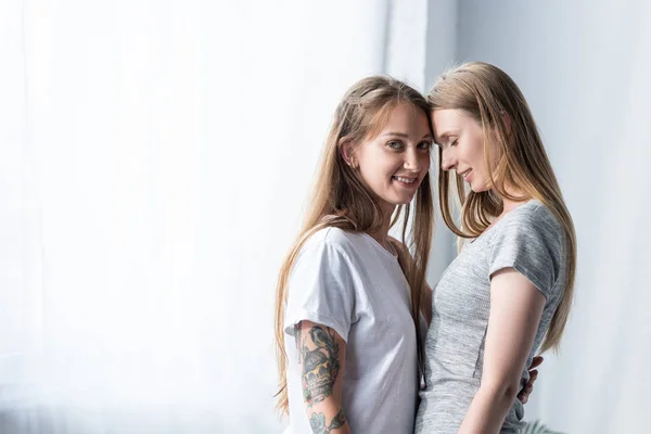 Deux lesbiennes souriantes en t-shirts embrassant dans la chambre — Photo de stock