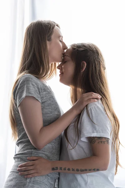 Vue latérale de lesbienne embrassant petite amie et l'embrassant dans le front à la maison — Photo de stock