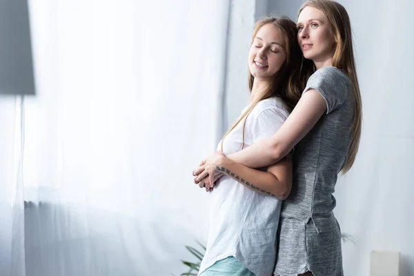 Дві усміхнені лесбіянки в футболках, що обіймаються в спальні — стокове фото