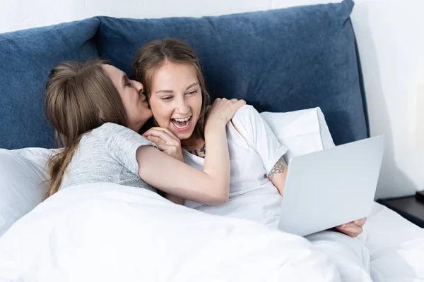 Dos lesbianas sonrientes abrazando y besándose mientras usa el portátil en la cama por la mañana - foto de stock