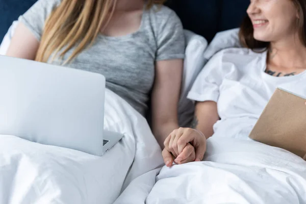 Vista parcial de dos lesbianas con portátil y libro cogidas de la mano mientras yacen debajo de la manta en la cama - foto de stock