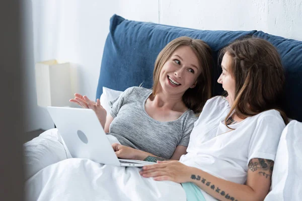 Dos lesbianas sonrientes mirándose mientras usan el portátil en la cama por la mañana - foto de stock