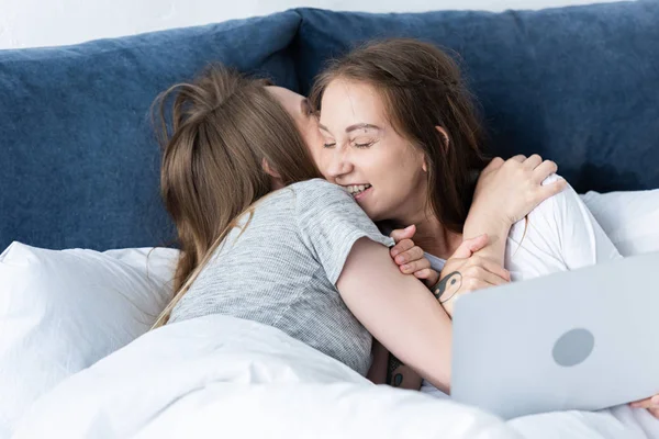 Due lesbiche sorridenti che si abbracciano mentre usano il computer portatile a letto al mattino — Foto stock