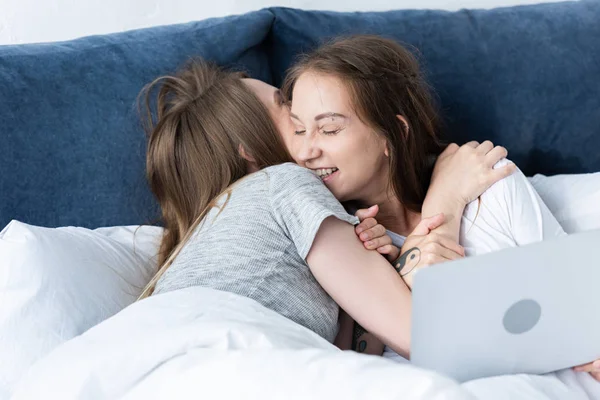 Duas lésbicas sorridentes abraçando ao usar laptop na cama de manhã — Fotografia de Stock