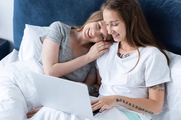 Zwei lächelnde Lesben, die sich umarmen, während sie morgens Laptop im Bett benutzen — Stockfoto