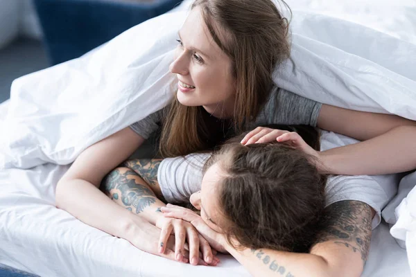 Дві усміхнені лесбіянки обіймаються, лежачи під ковдрою в ліжку — стокове фото