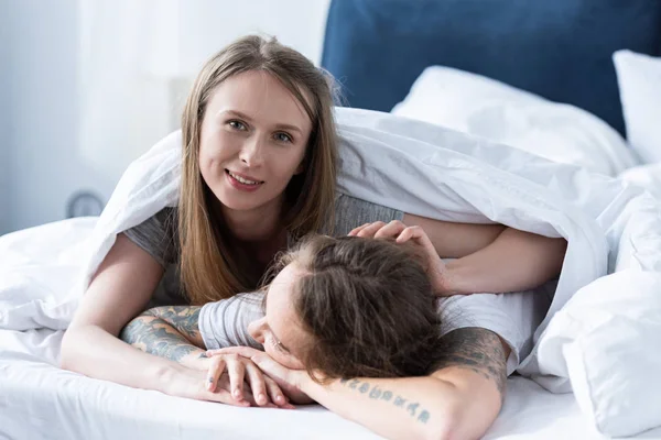 Zwei lächelnde Lesben umarmen sich, während sie unter einer Decke im Bett liegen — Stockfoto