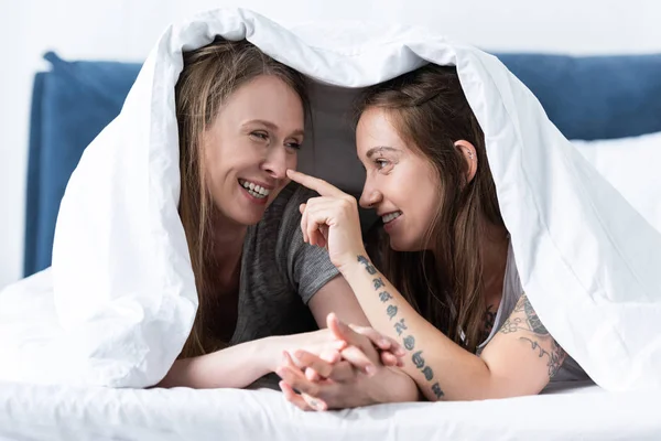 Two happy lesbians holding hands while lying under blanket on bed — Stock Photo