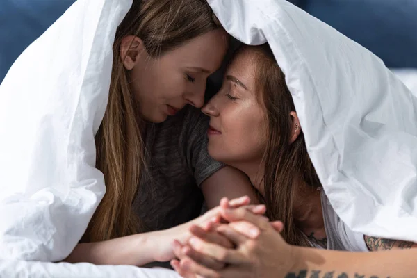 Two happy lesbians holding hands while lying under blanket on bed — Stock Photo